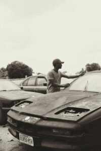 A man in a cap touches a vintage car in a parking lot with other vehicles.
